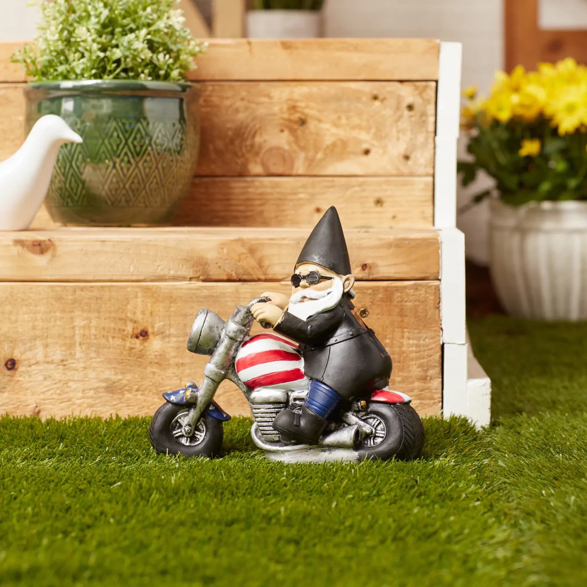 Gnome on a motorcycle with an American flag design, sitting on grass with wooden planters and flowers in the background.