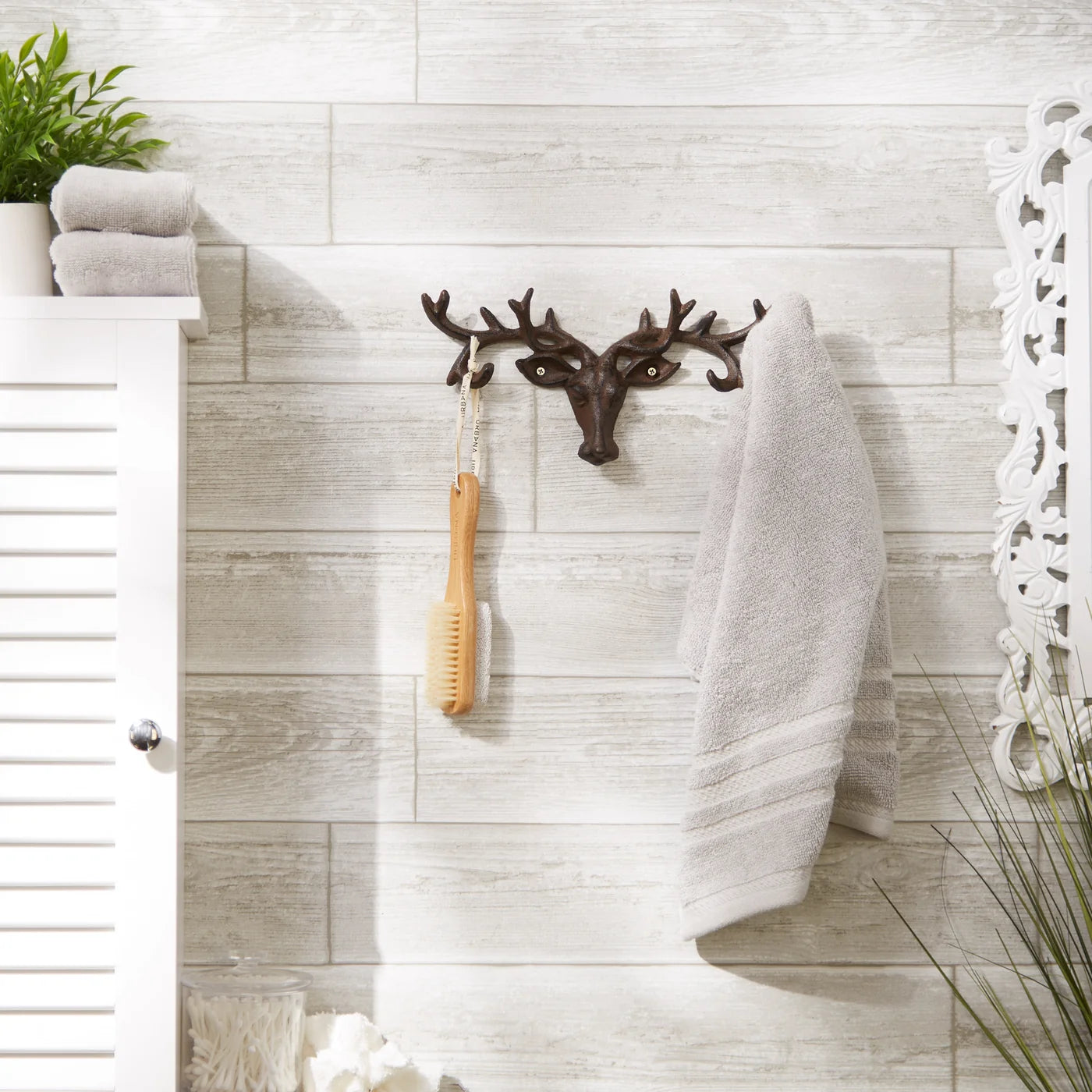 A dark brown metal deer head wall hook with antlers hangs on a light gray wood-paneled bathroom wall. A beige towel is draped over the right side of the hook, and a wooden brush hangs from the center.