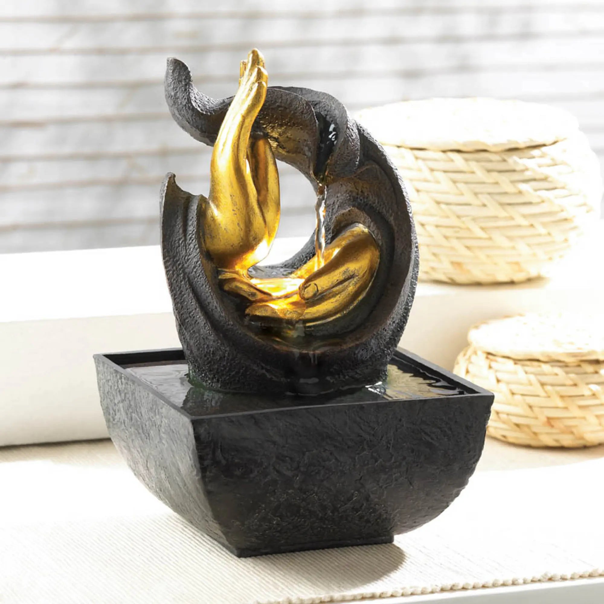 Decorative tabletop fountain with black and gold hands pouring water, placed on a surface with a blurred background.
