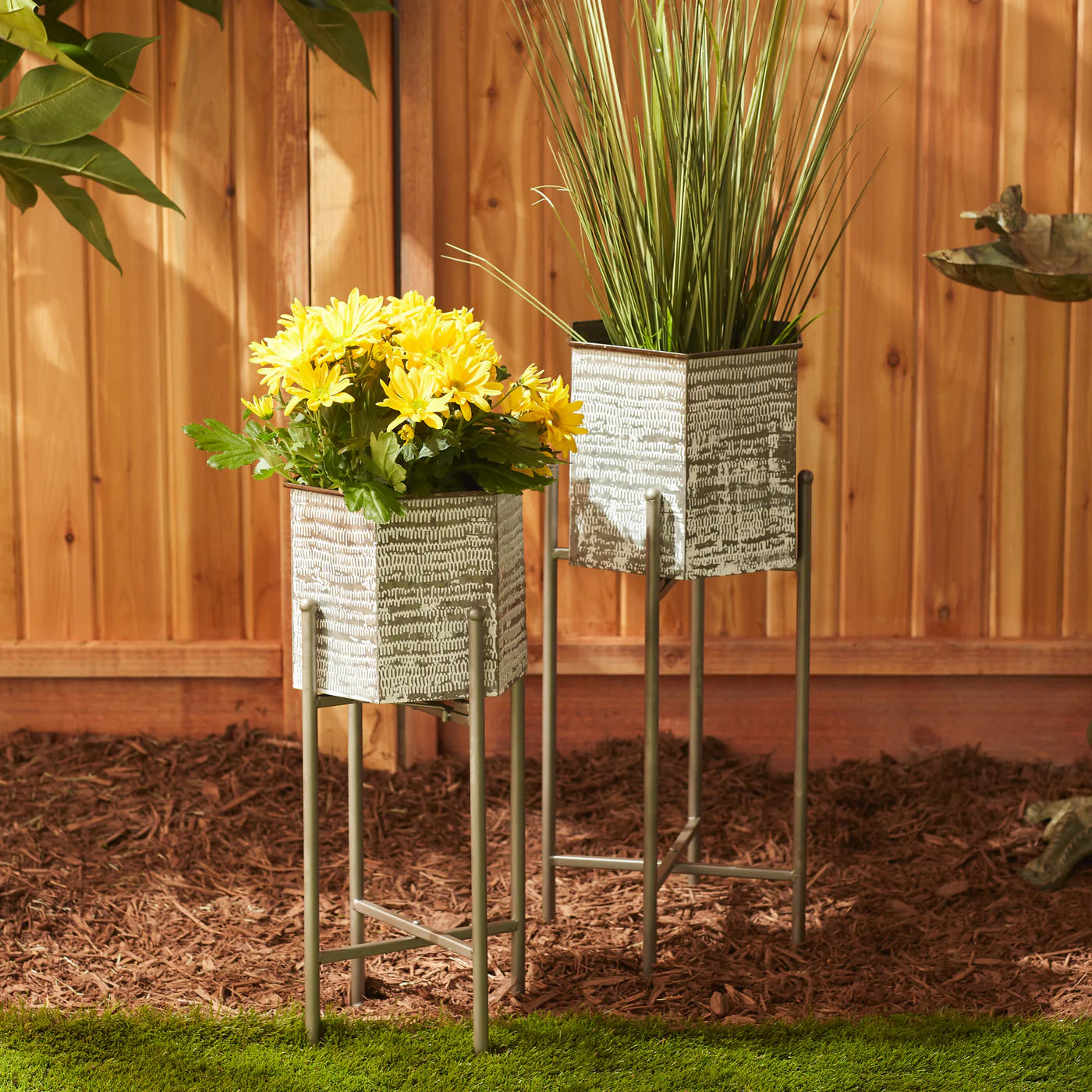 Two planters with yellow flowers and green plants on stands against a wooden fence.