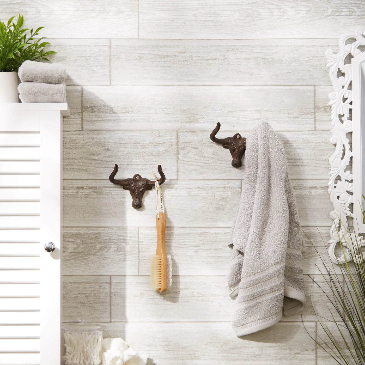 Set of 2 cast iron longhorn wall hooks holding a towel and bath brush on a tiled bathroom wall.