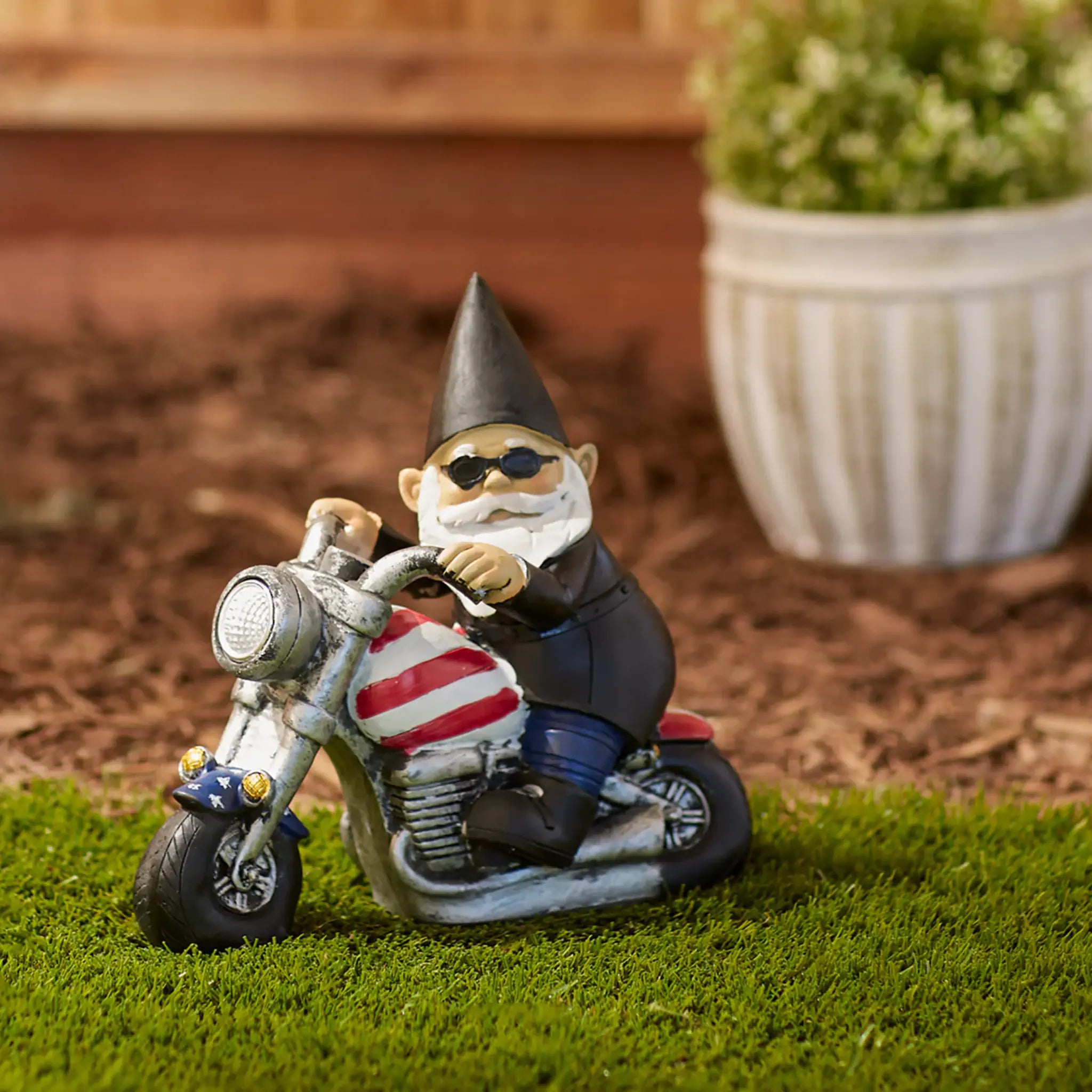 Gnome on a motorcycle with an American flag design, sitting on grass with a potted plant in the background.